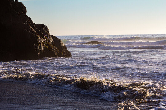 View of sunlight catching the crests of waves rolling onto a dark sandy beach, contrasting with the rugged, shadowed rocks, Fort Bragg, California, United States.