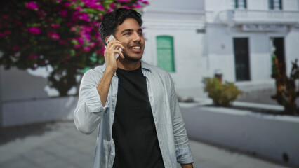 Man holds smartphone to ear smiling on a street beside a white building and blooming bougainvillea;...