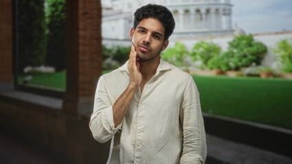 Fototapeta premium Man with hand on cheek and hand on chest over beige linen shirt in front of a building courtyard with lawn and dome; thoughtful introspection.