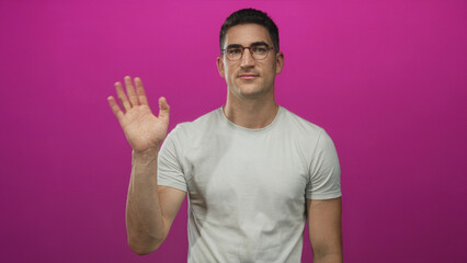 Naklejka premium Man wearing glasses and light t shirt waving right hand with palm visible in studio against bright magenta backdrop; friendly greeting.
