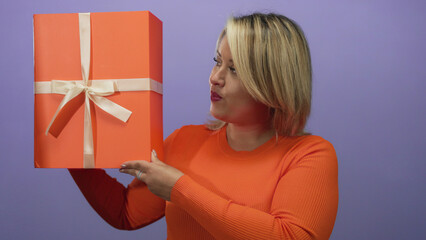 Woman with blonde hair holding large orange gift box with cream ribbon in purple studio;...