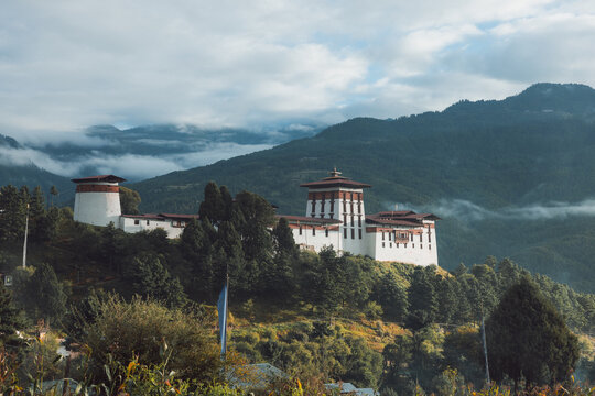 View of a majestic dzong perched atop a hill amidst lush greenery, with mist-shrouded mountains in the background, creating a serene atmosphere, Jakar, Bumthang, Bhutan.