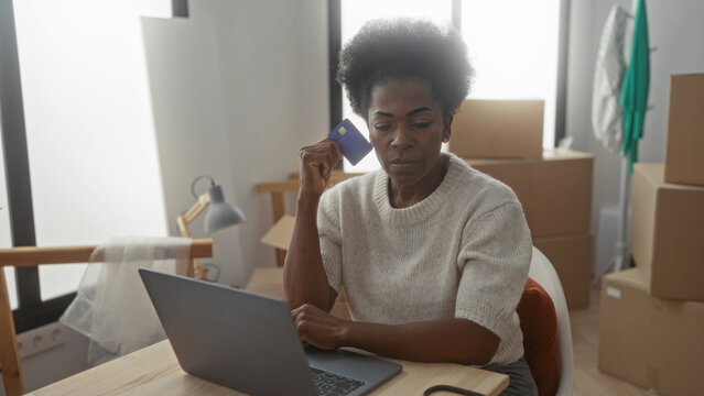 Woman using laptop in new apartment surrounded by moving boxes holding credit card looks thoughtful implying online shopping or financial planning