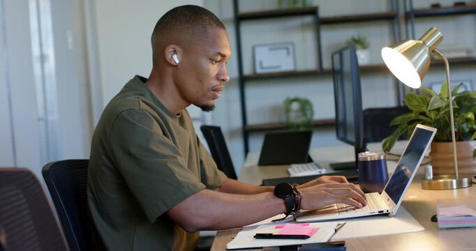 Man typing on laptop at office desk with earbuds and coffee mug pausing while acknowledging camera