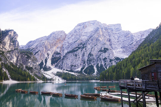 View of small wooden boats float on the tranquil turquoise lake beneath towering, snow-dusted mountains and dense green forests, Campitello di Fassa, Italy.