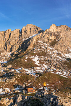 View of a lone hiker in a yellow jacket stands atop rocky outcrop, gazing at the majestic Dolomites mountains under a clear blue sky, Campitello di Fassa, Trentino-Alto Adige, Italy.