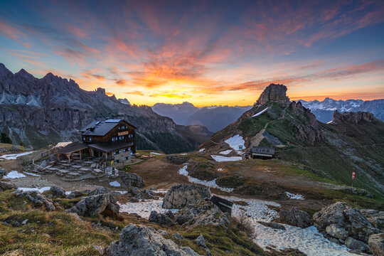View of a rustic mountain lodge nestled amidst jagged peaks under a vibrant sunset sky, where fiery hues paint the clouds in Campitello di Fassa, Trentino-Alto Adige, Italy.