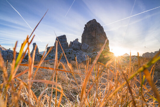 View of golden grasses sway in foreground as the sun kisses jagged peaks beneath a sky streaked with contrails, Dolomites, Campitello di Fassa, Trentino-Alto Adige, Italy.