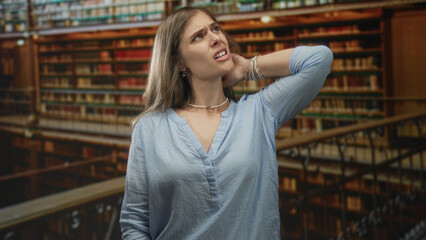 Fototapeta premium Blonde woman in blue blouse holds hand to neck amid wooden shelves in library building; discomfort.
