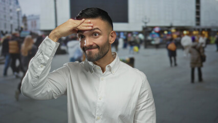 Obraz premium Young hispanic man on urban street outdoors looking thoughtful with hand on forehead as diverse city crowd blurs by in background.
