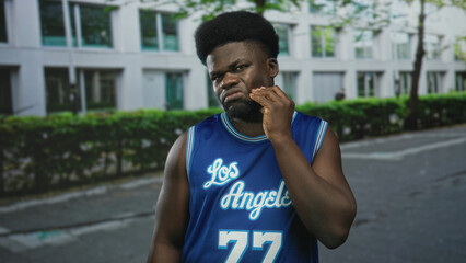 Man in blue basketball jersey with hand to cheek for toothache on city street near hedge;...