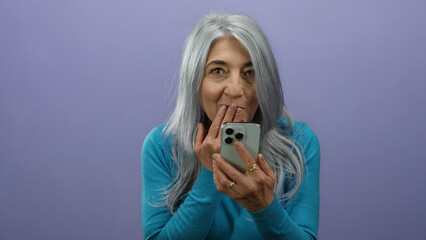 Fototapeta premium Senior woman with grey hair using smartphone against purple background, expressing surprise and amusement in isolated vibrant setting.