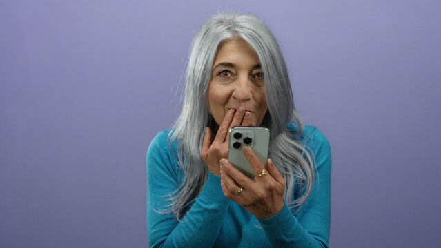 Senior woman with grey hair using smartphone against purple background, expressing surprise and amusement in isolated vibrant setting.