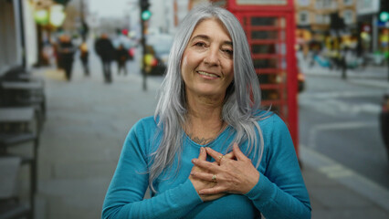 Fototapeta premium Senior woman with grey hair standing on a busy london street in front of a classic red phone booth, expressing emotion with her hands clasped to her chest.