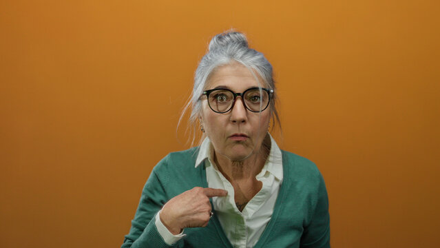 Woman with grey hair and glasses pointing at herself, wearing a green cardigan against an orange background, expressing surprise and curiosity.