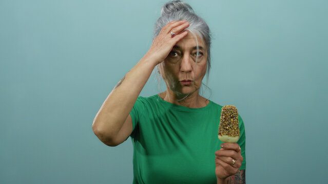 Elderly woman with grey hair in a green shirt holding an ice cream bar against a blue background, looking expressive and isolated with a surprised demeanor.