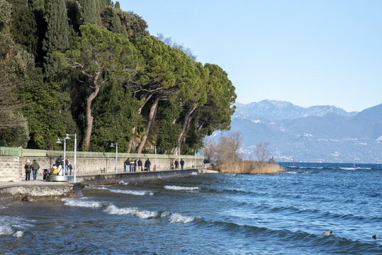 View of gentle waves lapping against the shore, people strolling along the promenade under the shade of towering trees, with snow-capped mountains, Sirmione, Lombardy, Italy.
