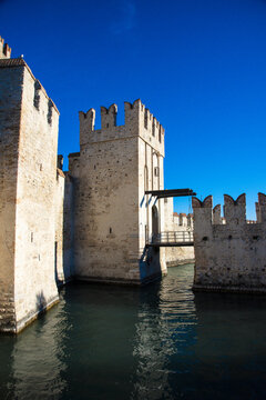 View of a medieval castle with imposing stone walls rises from the waters, a bridge connecting it under a vibrant blue sky, Sirmione, Lombardy, Italy.