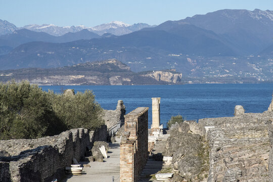 View of ancient ruins with exposed brick and stone walls leading towards the brilliant blue waters of a lake, framed by distant mountains, Sirmione, Lombardy, Italy.