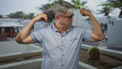 Senior man with grey hair and glasses flexes biceps, rubs elbow and grimaces while posing on a sunny street; aging pride.