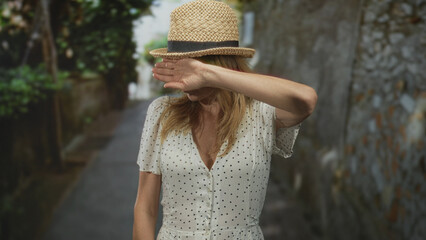 Woman shields face with hand on narrow street wearing straw hat and white polka dot dress; privacy annoyance.