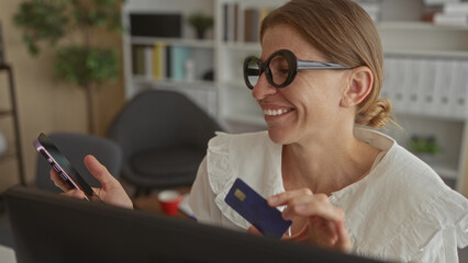 Woman smiling while holding creditcard and smartphone at office desk, wearing round glasses and gesturing to the card; online payment joy.