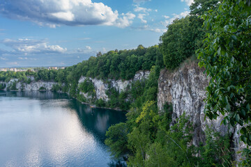 Obraz premium A calm blue quarry lake surrounded by tall limestone cliffs and dense green forest under a cloudy sky. The scene represents untouched nature, dramatic geology, outdoor recreation, and peaceful landsca