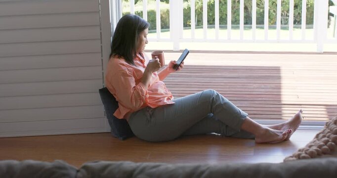 Woman is sipping mug and reading phone by deck as sunlight is drawing gaze, watching garden