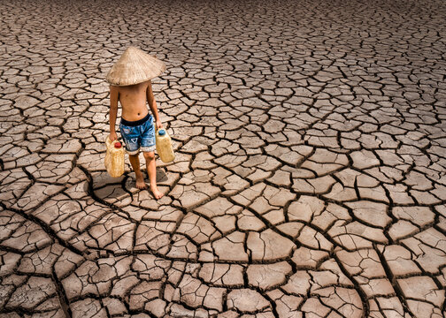 View of cracked earth under the harsh sun as a child carries water jugs in a desperate search for water, highlighting the drought's impact, Bac Lieu, Vietnam.