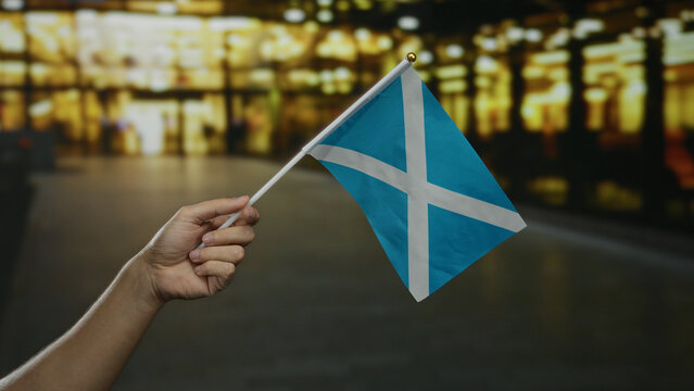 Hand holding scottish flag in city street showcasing national pride outdoors at night with blurred lights in the background.