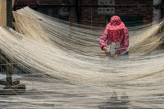 View of sun-drenched noodles cascade around a worker in a red floral hat, creating an ethereal veil in the open air, Taichung, Taichung, Taiwan.