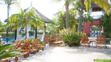 Fototapeta premium Blurred view of a tropical luxury resort with wooden lounge chairs, palm trees, and a serene poolside area bathed in sunlight, exuding tranquility and relaxation.