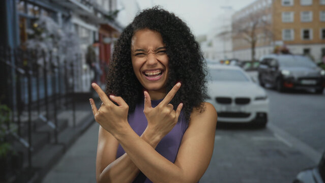 Woman smiling broadly showing rock horns gesture with both hands on bustling street lined with buildings; fun energy.