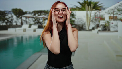 Fototapeta premium Woman smiling joyfully by a luxurious hotel pool in an outdoor resort setting, showcasing red hair, glasses, and casual attire with a modern and refreshing ambiance.