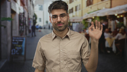 Obraz premium Young hispanic man with glasses waves hand in casual shirt on street terrace outdoors; friendly greeting.