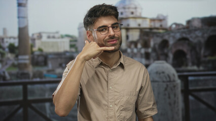 Hispanic man wearing eyeglasses and beige shirt making phone gesture in front of ancient roman building ruins; connection.
