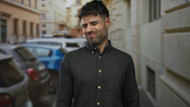 Man in black buttondown shirt grimaces with tightly closed eyes and tense face on narrow cobbled city street; disgust.