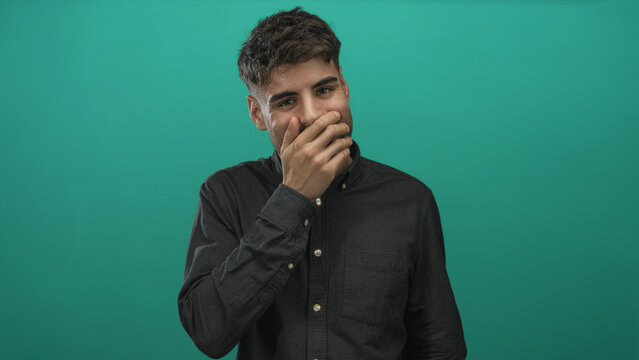 Young hispanic man covers mouth with hand and smiles shyly against green studio wall portrait; surprise.