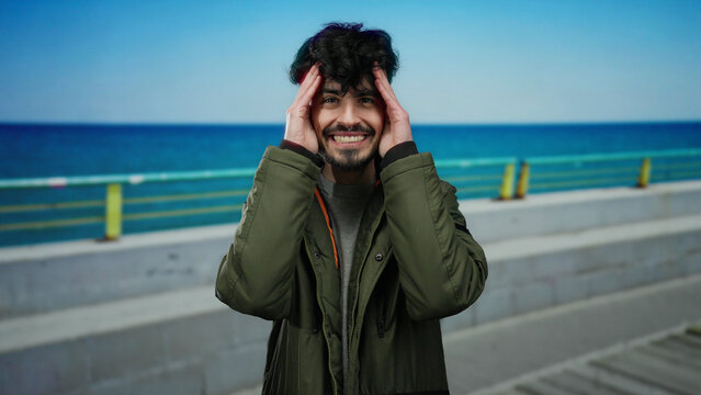 Handsome man standing on a seaside promenade with a jacket, expressing various emotions against the blue sea and sky backdrop, capturing a young adult enjoying the serene beachscape.