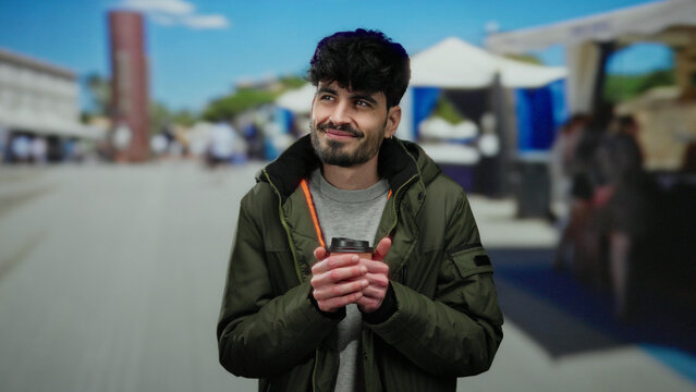 Young man smiling with coffee cup in hands standing at outdoor market street under a clear blue sky, wearing a green jacket, capturing a relaxed and happy moment in a vibrant urban setting.