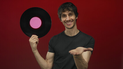 Young man smiling holds vinyl record by pink label on fingers in red studio; nostalgia music art...