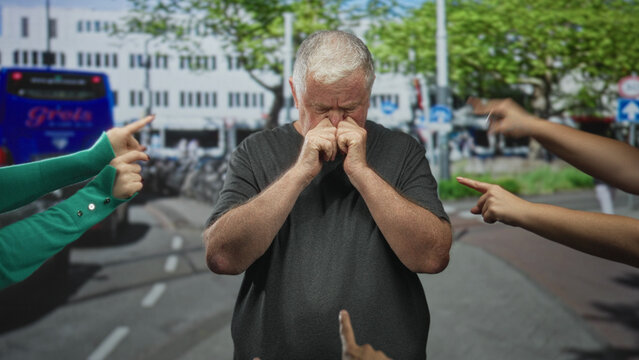 Man senior rubbing eyes with hands while several people point fingers at him on a busy street beside a bus and bicycle rack near buildings; humiliation isolation.