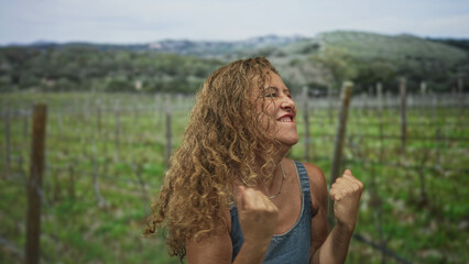 Obraz premium Woman clenches fists and smiles in denim dress amid a vineyard with wooden trellis in forest hills; joy triumph.