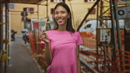 Fototapeta premium Thai young brunette woman holds toothbrush and smiles, wearing pink tshirt while standing amid scaffolding on street; cheerful oral care.