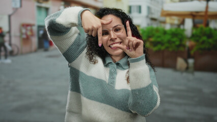 Fototapeta premium Young woman posing playfully on city street wearing glasses and striped sweater, creating frame with fingers during daytime, showing joyful expression in urban setting.