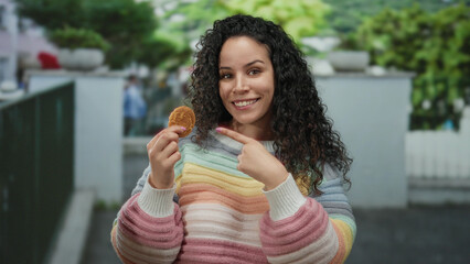 Fototapeta premium Woman smiling outdoors wearing colorful sweater holding cookie on street with trees in background, showcasing cheerful and relaxed mood in open urban environment.