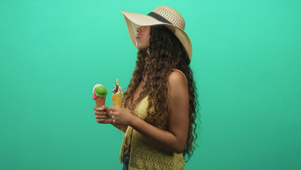 Woman holding two icecream cones, sunhat tilted, yellow crochet top and jeans in studio with green backdrop, neutral steady gaze; summer contemplation.