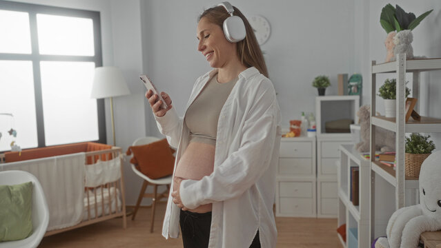 Pregnant woman listening to music with headphones and using phone in bright baby room, smiling near crib and shelves filled with toys, showcasing a cozy and modern home environment.