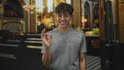Man in gray shirt with curly hair making ok sign with right hand and smiling in ornate church...