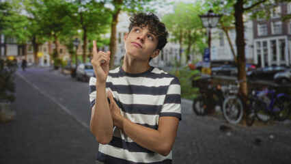 Teen boy in a striped shirt pointing finger upward with exposed forearm on a city street beside parked bicycle and lamppost; curiosity daydreaming imagination.